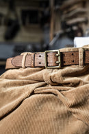 A close-up of a brown textured leather belt with a brass buckle, draped over a tan corduroy fabric in a softly blurred workshop setting, Obroi