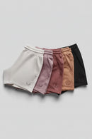 Five pairs of women's shorts in white, dusty pink, mauve, tan, and black, arranged in a fan shape on a light gray background. Each pair features a small embroidered logo on the lower left leg, Obroi