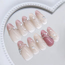 A set of artificial press-on nails arranged on a white heart-shaped tray. The nails feature a glossy gradient design transitioning from pale pink to soft nude beige, with white French tips, delicate butterfly wing patterns on some nails, and silver rhinestone and pearl embellishments, Obroi