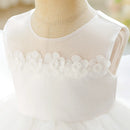 White sleeveless flower girl dress with a sheer mesh yoke and 3D fabric flower applique across the chest, displayed on a beige mannequin torso, Obroi