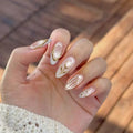 Hand with decorative nail art featuring gold and white designs against a blurred natural background, Obroi