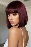 A woman with a short, straight bob wig in deep burgundy with straight-cut bangs, shown from the back over her shoulder in an indoor setting, Obroi