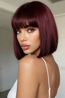 A woman with a short, straight bob wig in deep burgundy with straight-cut bangs, shown from the back over her shoulder in an indoor setting, Obroi