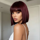A woman with a short, straight bob wig in deep burgundy with straight-cut bangs, shown from the back over her shoulder in an indoor setting, Obroi