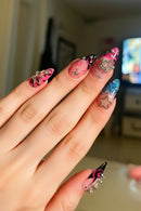 A close-up of hands showcasing long, glossy almond-shaped press-on nails with a variety of colorful designs, including pink, blue, black, and white patterns, decorated with metallic silver glitter stars and rhinestones, Obroi
