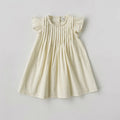 A cream-colored baby dress with ruffled cap sleeves and vertical pleats on the bodice, laid flat against a light gray background, Obroi