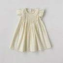 A cream-colored baby dress with ruffled cap sleeves and vertical pleats on the bodice, laid flat against a light gray background, Obroi
