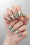 A close-up of manicured fingering with long almond-shaped nails decorated in a coordinated pale sage green and white set with metallic gold glitter borders, Obroi