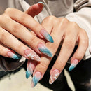Close-up of manicured fingernails on a person's hands, featuring almond-shaped tips with a blue-to-pink ombre gradient, white pearl accents, and two small white flower designs with gold centers, Obroi