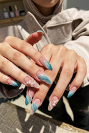 Close-up of manicured fingernails on a person's hands, featuring almond-shaped tips with a blue-to-pink ombre gradient, white pearl accents, and two small white flower designs with gold centers, Obroi