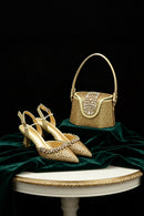 A pair of gold pointed-toe high heels with crystal embellishments is displayed next to a matching gold handbag with a crystal-embellished flap, set on draped dark green velvet against a black background, Obroi