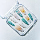 A set of 10 press-on acrylic nails displayed inside a clear plastic case on a textured white surface. The nails feature a mix of designs including teal and white marbled patterns, white seashells with pearl accents, and two white nails decorated with yellow tropical flowers, Obroi