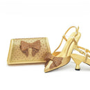 A gold metallic shoe and matching clutch set, both covered in rhinestones and featuring a large bow detail on the front, displayed on a clean white background, Obroi