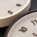 Close-up of a round wall clock with a textured light beige stone or concrete face, dark brown numerals, and a dark brown rim, shown against a dark background, Obroi