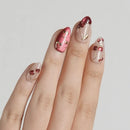 A close-up of a hand showcasing elegant almond-shaped nail art. The design features a glossy pink accent nail with gold cherry details, a nude clear nail with a cherry motif, and a metallic rose gold nail with red jewel accents, all set against a clean light gray background, Obroi