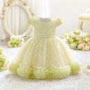 A pale yellow-green toddler girl's party dress with a beaded bodice, short puff sleeves, and a tiered tulle skirt with ruffled trim, displayed in a styled room setting with a mirror and flowers, Obroi