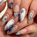 A close-up of a hand showcasing a detailed nail art design. The manicure features almond-shaped nails with a white base decorated with blue floral motifs, delicate gold metallic borders, and small clear rhinestone accents, Obroi