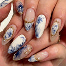 A close-up of a hand showcasing a detailed nail art design. The manicure features almond-shaped nails with a white base decorated with blue floral motifs, delicate gold metallic borders, and small clear rhinestone accents, Obroi