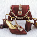 A coordinated women's fashion set featuring a structured burgundy handbag with a gold chain strap and ornate jeweled embellishment, paired with matching pointed-toe heeled sandals in burgundy satin with gold insoles and matching decorative embellishments on the toes, Obroi