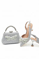 Silver clutch bag and high-heeled shoes with floral embellishments on a white background, Obroi