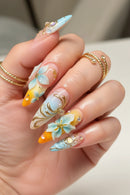 A close-up of a hand showcasing elaborate, long almond-shaped nails with pastel blue, yellow, and white gradient designs, gold metallic swirls, 3D floral accents, and small pearl details. The hand is also wearing two gold rings, Obroi