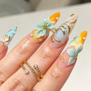 A close-up of a hand showcasing elaborate, long almond-shaped nails with pastel blue, yellow, and white gradient designs, gold metallic swirls, 3D floral accents, and small pearl details. The hand is also wearing two gold rings, Obroi