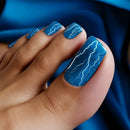 Nail art with blue polish and white lightning designs on a blue fabric background, Obroi