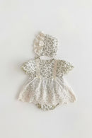 A baby girl's short-sleeve lace romper dress with a matching ruffled bonnet, featuring a delicate green and white floral print on a cream background, laid flat on a light gray surface, Obroi
