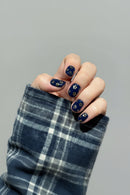 Hand with dark blue nail polish featuring floral designs, wearing a plaid sleeve against a gray background, Obroi