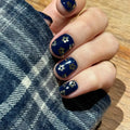 Hand with blue nail polish featuring gold floral designs on a plaid fabric background, Obroi