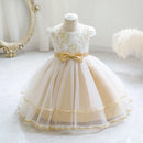 A toddler girl's formal party dress displayed on a mannequin. The dress features a white lace bodice with gold floral embroidery, a beige tulle skirt, and a matching beige bow at the waist, Obroi