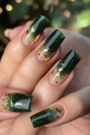 Three manicured fingernails shown on a hand, featuring a glossy dark green ombre top with a sheer nude-pink base and scattered metallic gold leaf accents, Obroi