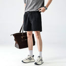 Person wearing black shorts and white sneakers holding a brown bag on a light gray background, Obroi
