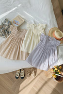 Three sleeveless children's dresses laid out on a white bed: one beige, one cream, and one light lavender, all with ruffled shoulders and vertical pleating. A floral scarf, book, straw hat, shoes, basket, and yellow flowers are arranged around them, Obroi