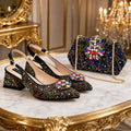 A pair of black pointed-toe slingback heels and a matching embellished handbag displayed on a marble table with ornate gold detailing. The shoes and bag are covered in multicolored rhinestones, with floral embellishments on the front of the shoes and a large floral brooch on the bag, Obroi