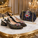 A pair of black pointed-toe slingback heels and a matching embellished handbag displayed on a marble table with ornate gold detailing. The shoes and bag are covered in multicolored rhinestones, with floral embellishments on the front of the shoes and a large floral brooch on the bag, Obroi