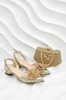 A pair of gold glitter slingback heeled sandals with large 3D fabric flower embellishments on the toes, shown alongside a matching gold clutch handbag with a chain strap, set against a white background with wavy lines, Obroi