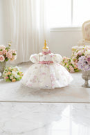 A baby girl's white tulle dress with pink and green floral prints and butterfly motifs, displayed on a wooden dress form mannequin. The dress features puff sleeves, a pink satin waistband with a bow, and a pink bow at the collar, Obroi