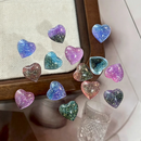 A group of small heart-shaped resin cabochons with glittery galaxy patterns in various colors, arranged on a light beige textured mat with a wooden frame and a glass tumbler in the background, Obroi