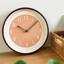 A round wall clock with a warm peach dial, white inner ring, dark brown outer rim, and dark brown hands, shown resting on a wooden surface against a light wall with a woven basket nearby, Obroi