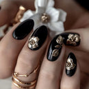 A close-up of manicured fingernails with a glossy black base and metallic rose-gold floral and leaf designs, shown on a hand with rose-gold rings and a blurred white flower in the background, Obroi