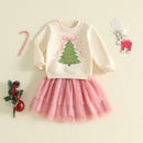 A children's holiday outfit laid flat on a light cream background, featuring a cream sweatshirt with a green Christmas tree and pink bow graphic, paired with a pink tulle skirt with small blue and gold star details. Decorative holiday props including a candy cane, red ornament, pine sprig, snowflake, and reindeer are arranged around the outfit, Obroi