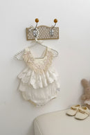 White lace baby romper on a hanger with baby shoes and a plush toy on a light surface, Obroi
