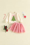 A toddler girl's Christmas-themed two-piece outfit featuring a cream sweatshirt with a green Christmas tree graphic and a pink tulle skirt with silver glitter, styled on a light beige background with holiday props, Obroi