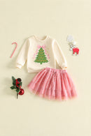 A toddler girl's Christmas-themed two-piece outfit featuring a cream sweatshirt with a green Christmas tree graphic and a pink tulle skirt with silver glitter, styled on a light beige background with holiday props, Obroi