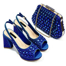 A matching blue evening wear set including a pair of open-toe high-heeled sandals and a small clutch purse, both embellished with blue rhinestones and gold-tone hardware, on a white background, Obroi