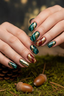 Close-up of hands with festive nail art featuring gold and red designs, set against a natural background with pine cones and acorns, Obroi