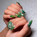 Close-up of a hand with green and white floral nail art on a textured gray background, Obroi