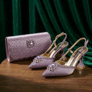 A pair of lavender satin slingback high heels with crystal embellishments and gold heels, displayed next to a matching lavender clutch bag on a wooden table with a dark green velvet curtain background, Obroi