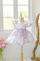 A toddler girl's white dress with a purple lavender floral print, puff sleeves, and a matching purple ribbon bow at the waist, displayed on a wooden dress form in a bright room, Obroi
